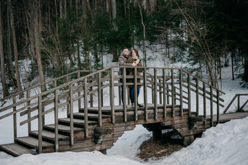 Stylish strong man with his cute young wife embrace in the forest on the beautiful old wooden bridge.