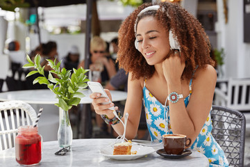 Sideways shot of attractive curly woman has happy expression, enjoys electronic song in modern headphones, has recreation time, reads text message on cell phone, poses outdoors with dessert.