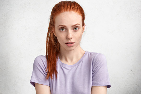 Isolated studio shot of beutiful young woman has freckled skin, red hair, wears casual purple t shirt, looks seriously at camera, listens interlocutor attentively, poses against white concrete wall