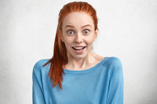 Portrait of astir overjoyed beautiful female, has green eyes, ginger hair and freckled skin, feels suprisment and excitment, recieves propoasal from boyfriend, isolated over white concrete wall