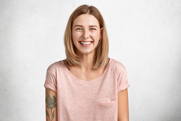 Happy delighted female with positive smile, has tattoo, smiles broadly, dressed in casual clothing,...