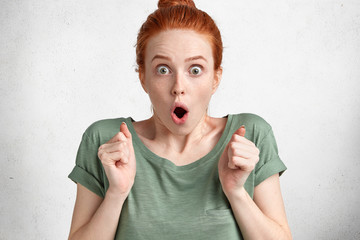 Shocked stunned frightened freckled ginger young female, stares at camera and has unexpected reaction on shocking news, keeps hands in fists, isolated over white studio background. Omg concept - obrazy, fototapety, plakaty