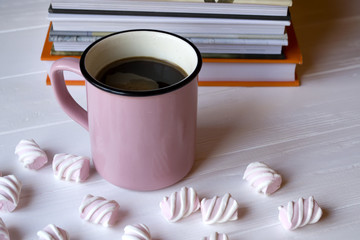 The pink mug of hot coffee with marshmallow and books on a white wooden table.