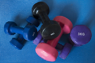 A pile of neoprene coated iron dumbbells of different weights, used for muscle toning, aerobic and weight training, positioned on a blue mat, part of fitness classes and at-home workout routine