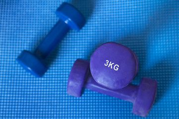 Neoprene coated iron dumbbells, a pair of 3 kg and a single of 2 kg, used for muscle toning, aerobic and weight training, positioned on a blue mat, part of fitness classes and at-home workout routine