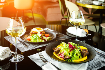Romantic dinner at a restaurant. Appetizing dishes with meat and lettuce leaves and glasses with wine in rays of the sun