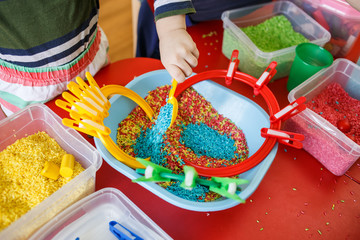 Toddlers playing with sensory bin with colourful rice on red table. - obrazy, fototapety, plakaty