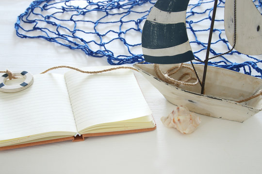 nautical concept image with white decorative sail boat and empty open notebook over white wooden table.