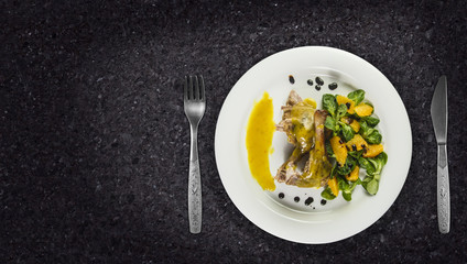 Duck legs with orange sauce and lamb's lettuce salad with oranges and vinegar on white plate on granite counter top.