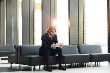 Young busy broker with tablet sitting on black leather sofa in airport lounge and networking while waiting for his flight