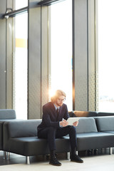 Young elegant businessman in suit sitting on leather sofa of airport lounge and searching for information for new project in the net