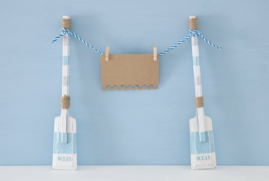 nautical concept with wooden decorative boat oars and empty note for copy space hanging on a string over blue background.