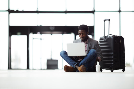 Cross-legged businessman with laptop sitting on the floor in airport lounge with suitcase near by and watching online video or chatting to someone