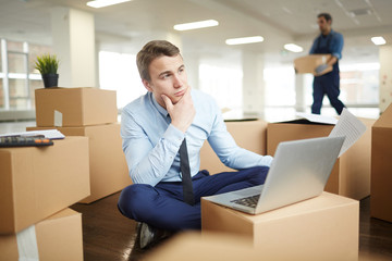Pensive businessman with paper sitting on the floor in front of laptop on top of packed box in new office
