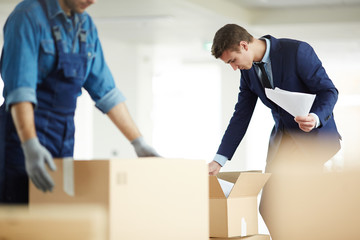 Contemporary businessman in suit looking for necessary papers in one of boxes just after relocation to new office