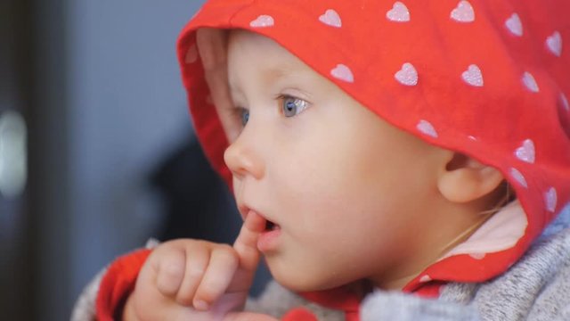 Close-up shot of blue eyed baby face watching cartoons. The child in red hood with silver hearts pattern sucks his finger looking with interest what happening on the screen