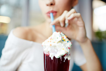 Glamorous girl drinking tasty cocktail with sprinkled ice-cream top through straw while relaxing in cafe