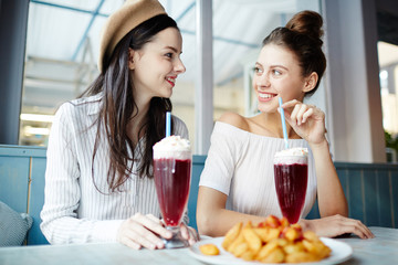 Happy young women sitting in cafe and enjoying taste of dessert or cocktail and fast-food