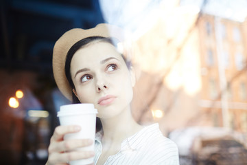 Pretty young woman in beige beret having drink in cafe and looking through window at urban scene
