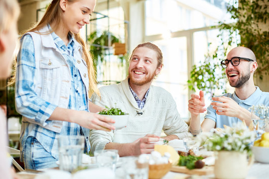 Two happy guys looking at young woman with bowl of vegetable salad prepared for dinner