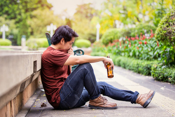 Sad single asian man drinking beer and sitting on the floor at public park.