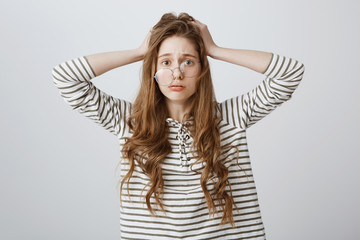 Fototapeta premium Girl feeling frustration from inability to fix problem. Portrait of worried and stressed young woman holding hands on hair, staring through glasses with nervous and upset expression over gray wall