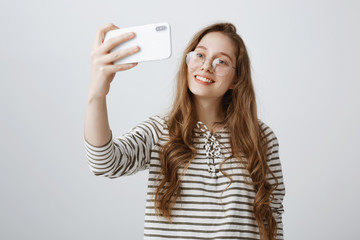 Fototapeta premium Modern teenagers cannot spend time without smartphones. Portrait of attractive urban girl taking selfie on phone, smiling and expressing positive emotions, standing over gray background