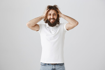 Headache mixed up thoughts. Stressed miserable eastern man with curly hair and beard holding head and grimacing from pain and negative emotions, being sad and devastated over gray background