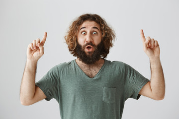 Guess what I saw upwards. Portrait of shocked and thrilled adult guy with beard and curly hair pointing up with raised index fingers, gasping and saying wow, popping eyes over gray background