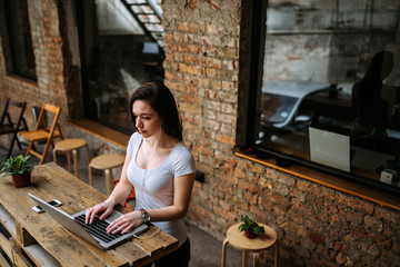 Lovely student girl using laptop and earphones while standing. Brick wall in the background.