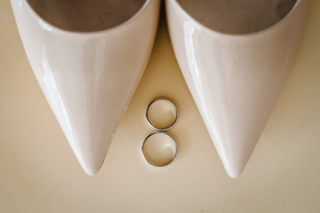 Stylish elegant classic lacquered beige shoes are isolated on table, two silver wedding rings lying on pastel background.  Wedding accessory standing. Close up. flat lay. top view.
