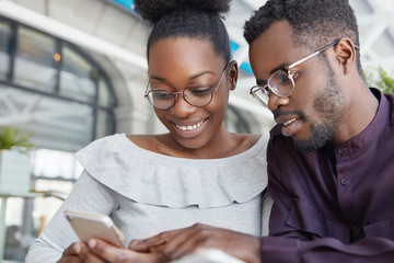 Photo of happy dark skinned African American friends, meet together, holds cell phone and watch photos or advertising video, wear spectacles, have pleasant smiles on faces. Ethnicity, technology