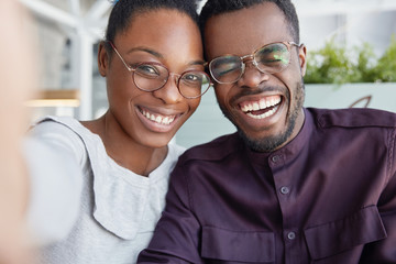 Positive dark skinned smiling woman and man have fun together, make selfie, wear glasses, laugh,...