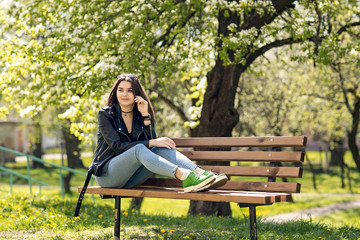 Glamorous young Caucasian woman in black leather jacket sitting in the park on the bench