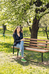 Obraz premium Glamorous young Caucasian woman in black leather jacket sitting in the park on the bench
