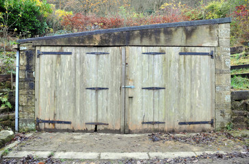 small old stone storage or garage building with decaying wooden doors and rusted hinges with damp walls and roof in a rural setting