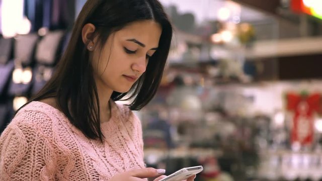 Profile of a stylish and nice girl touching the screen of her smartphone and estimating prices  while standing in a glassy shopping mall