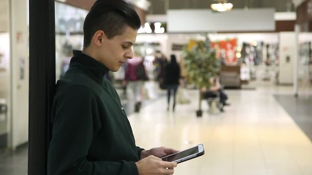 Profile of a slender  young man with crew haircut who stands, touches the screen and looks at his computer to analyze the prices in a mall