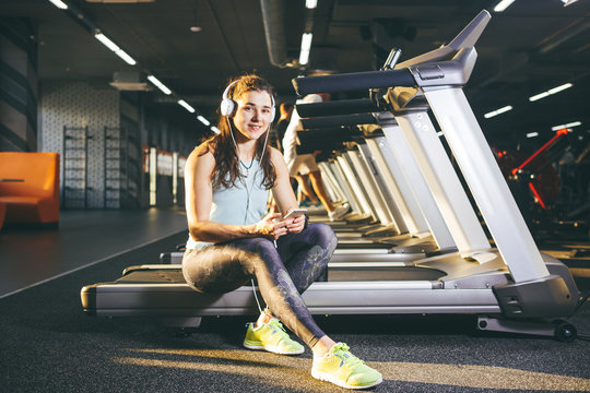 Beautiful young Caucasian girl sportswoman sitting, resting after training on treadmill against the backdrop of gym in sunny weather. Listens to music in sick white headphones,in hand holds the phone