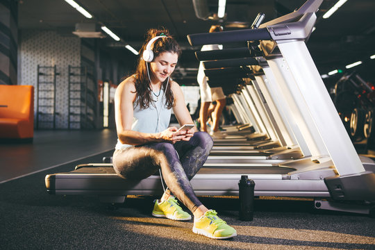 Beautiful young Caucasian girl sportswoman sitting, resting after training on treadmill against the backdrop of gym in sunny weather. Listens to music in sick white headphones,in hand holds the phone