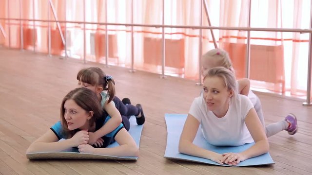 Mom and daughter are resting in the gym. The daughter lies on her mother's back and rests after sports exercises, she tells her secrets in her ear.