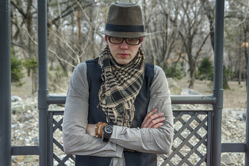 Stylish man in hat, scarf, waistcoat and shirt sitting on a bench in park, crossed his arms with vintache leather bracelets over his chest