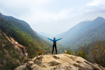 Tourist traveler standing on a rock with raised hands, hiker looking to a valley below in trip in...