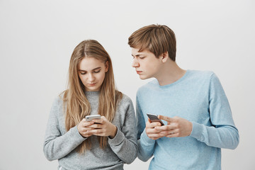 Two smartphone geeks posting new photos in social network. Portrait of focused cute friends with fair hair, holding cellphones and messaging, while guy peeking at girls screen to check what she writes