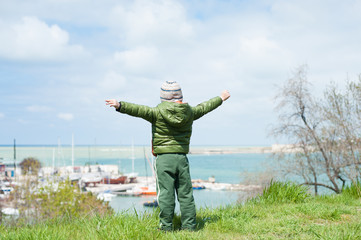 happy little boy in jacket and hat stands on green hill on background of sea bay with ships in early spring