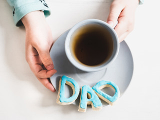Cup of fragrant tea and biscuits in the form of letters D A D in the hands of the younger daughter on a white background. View from above. Congratulations for daddy