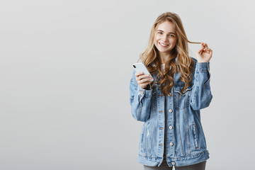 Can you add me to your friend list. Portrait of cute fashionable woman in denim jacket, playing with strand of hair while holding smartphone and smiling broadly, flirting with coworker over gray wall