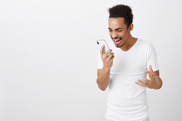 What the hell going on. Portrait of shocked outraged young dark-skinned guy with afro haircut holding smartphone, shouting at screen, gesturing, being annoyed and angry while arguing over gray wall
