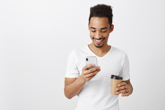 Sending Funny Meme To Girlfriend. Portrait Of Handsome African Coworker In Casual White Shirt, Holding Cup With Coffee And Smartphone, Typing Message Or Playing Game, Having Fun Over Gray Background