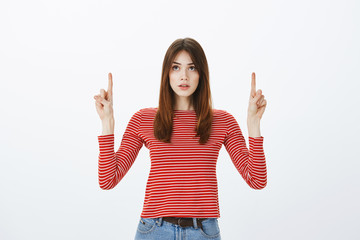 Studio shot of attractive woman feeling perplexed while thinking and focusing on item, hesitating buy it or not, raising index fingers, looking and pointing up, doubting and focusing on thoughts